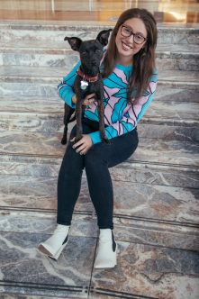 Jamie and her dog Lucy, a black dog with a red collar, are sitting on multicolored marble stairs. Jamie is wearing a light blue & pink patterned shirt with black pants and white boots. She is holding Lucy on right hand side.